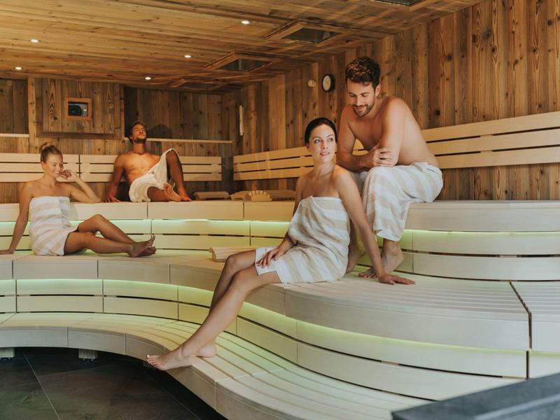 Vier Personen entspannen in einer hellen, modernen Sauna mit hellem Holz und weißen Bänken.