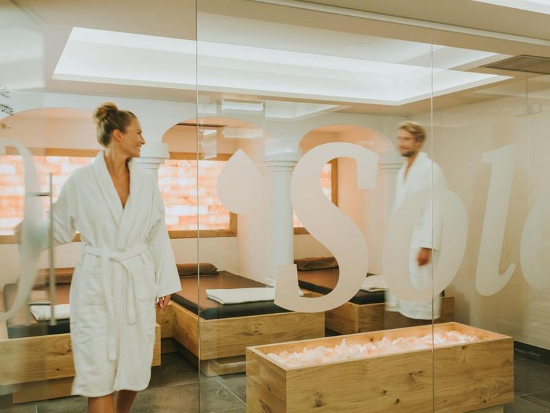 Zwei Personen in weißen Bademänteln stehen in einem hellen, modernen Spa-Raum mit Holzmöbeln.