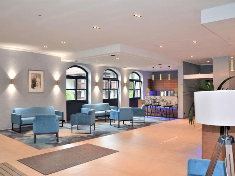 Nowoczesna lobby hotelowa z niebieskimi meblami, dywanami i dużymi oknami w jasnym pomieszczeniu.