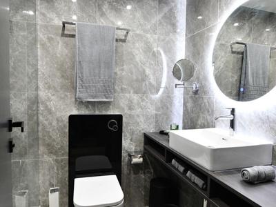 Modernes Badezimmer mit rundem beleuchtetem Spiegel, grauen Fliesen und weißer Toilette.