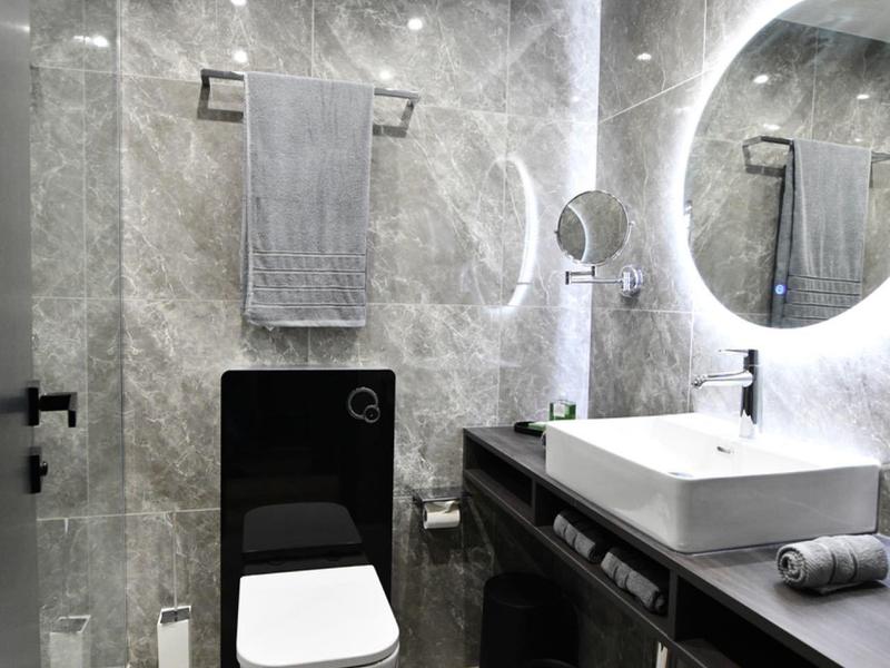 Baño moderno con azulejos grises, espejo iluminado y lavabo blanco sobre mueble oscuro.