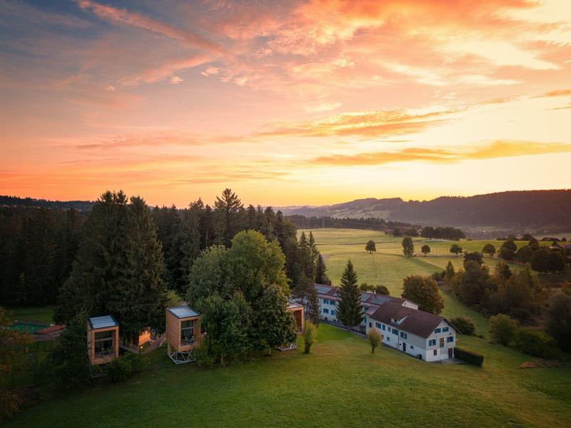 Landschaft mit Bäumen, mehreren kleinen Häusern und einem pastellfarbenen Sonnenuntergang.