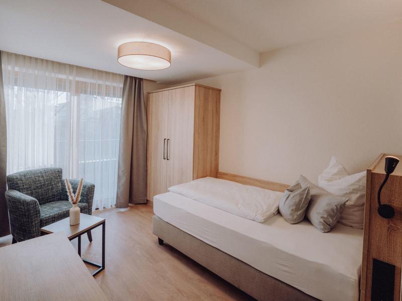 Jasny pokój hotelowy z pojedynczym łóżkiem, drewnianymi meblami i fotelem przy oknie