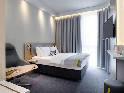 Modern eingerichtetes Hotelzimmer mit Doppelbett, Fernseher und großen Fenstern mit Vorhängen.