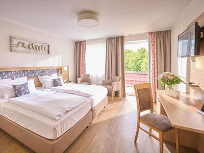 Helles Hotelzimmer mit Doppelbett, Schreibtisch, Fernseher und großem Fenster mit Blick ins Grüne.