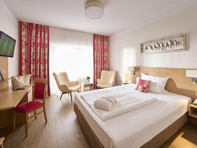 Helles Hotelzimmer mit Doppelbett, rotem Vorhang, Stühlen und Schreibtisch mit Fernseher