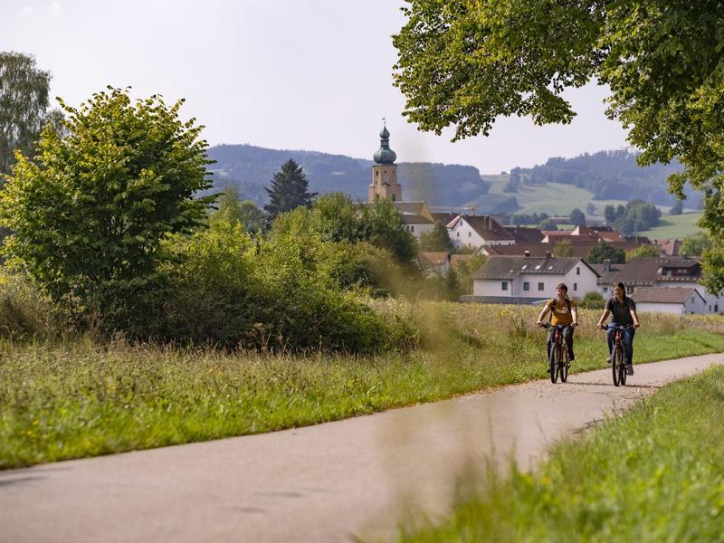 Due ciclisti percorrono un sentiero asfaltato accanto a campi verdi verso un villaggio con una torre campanaria.