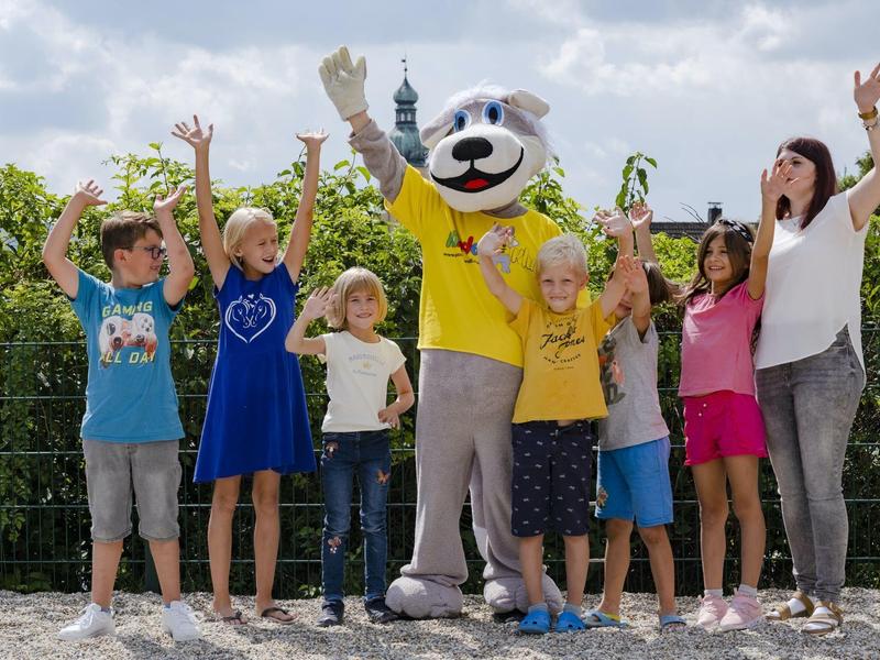 Gruppo di bambini entusiasti e un adulto che posano con una mascotte all'aperto in una giornata soleggiata.