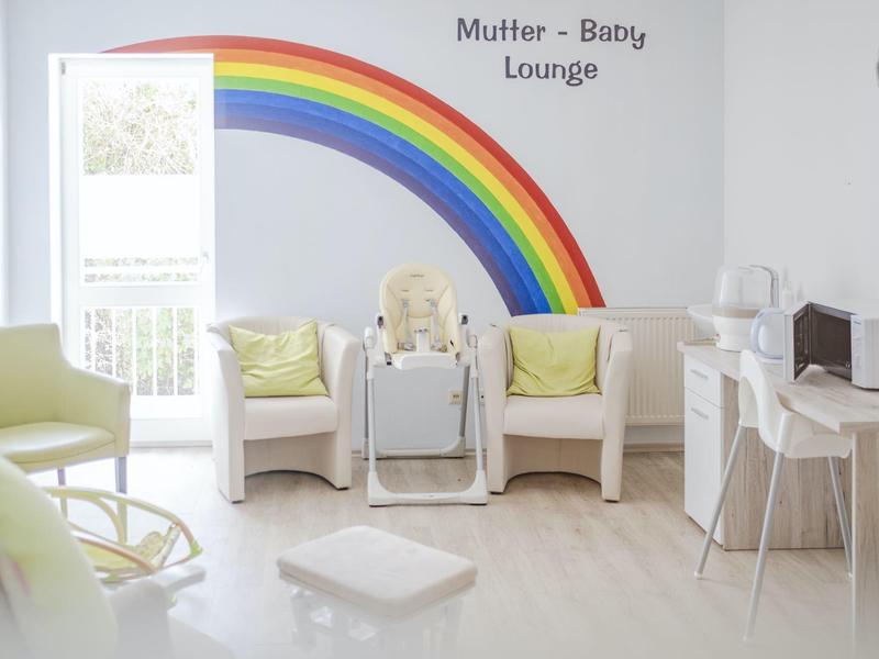 Sala luminosa con poltrone pastello, seggiolone e un arcobaleno adesivo con scritto Sala Madre-Bambino.