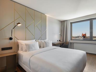 Helles Hotelzimmer mit Doppelbett, modernem Kopfteil und Fenster mit Blick auf Stadt