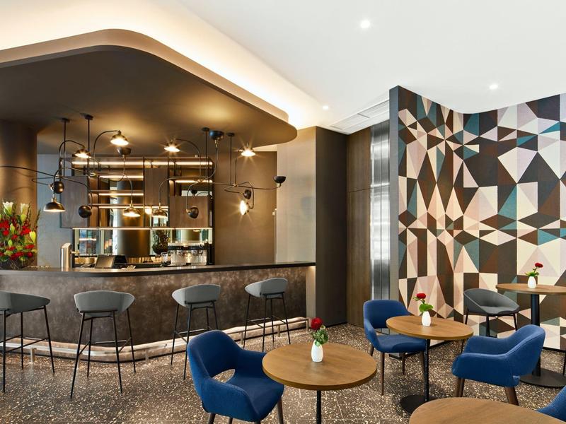 Nowoczesny bar hotelowy z geometryczną ścianą, ladą barową i niebieskimi fotelami przy drewnianych i marmurowych stolikach.