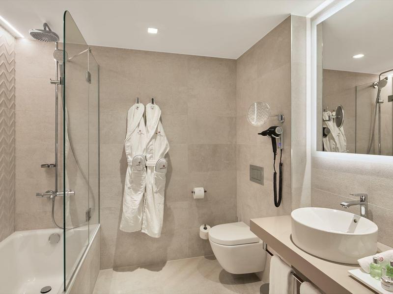 Nowoczesna łazienka hotelowa z wanną, dużym lustrem, umywalką i szlafrokami na ścianie.