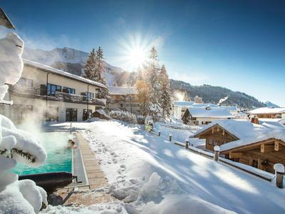 Winterlandschaft im Dorf mit verschneiten Holzhäusern, Sonnenschein und dampfendem Außenpool.