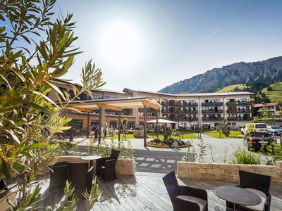 Außenbereich mit Tischen und Stühlen vor einem großen Hotelgebäude in sonniger Berglandschaft.