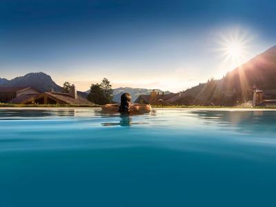 Frau schwimmt im Pool mit Bergkulisse im Sonnenuntergang, klares Wasser, heller Himmel.
