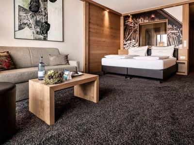 Modernes Hotelzimmer mit großem Bett, grauem Teppich, Sofa, Holzmöbeln und einem Kunstbild an der Wand.