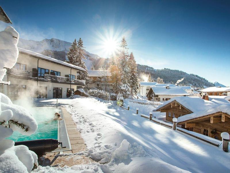 Winterlandschaft im Dorf mit verschneiten Holzhäusern, Sonnenschein und dampfendem Außenpool.