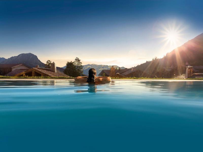 Frau schwimmt im Pool mit Bergkulisse im Sonnenuntergang, klares Wasser, heller Himmel.