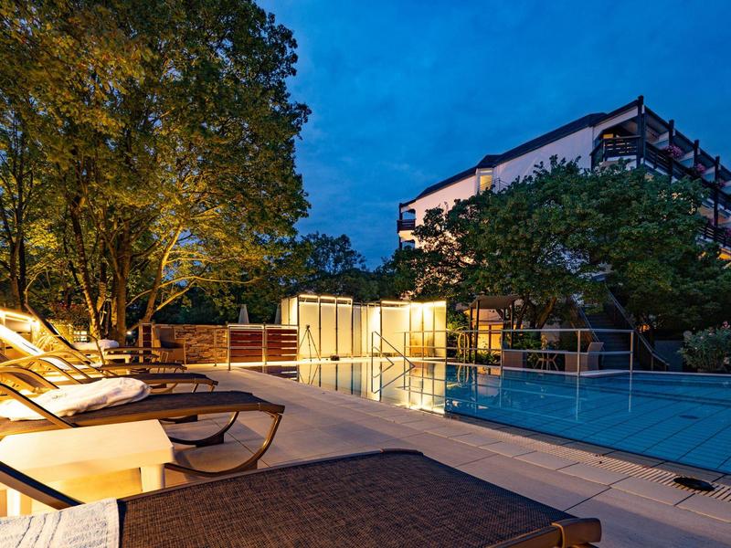 Piscina dell'hotel illuminata con lettini, alberi e cielo serale blu.