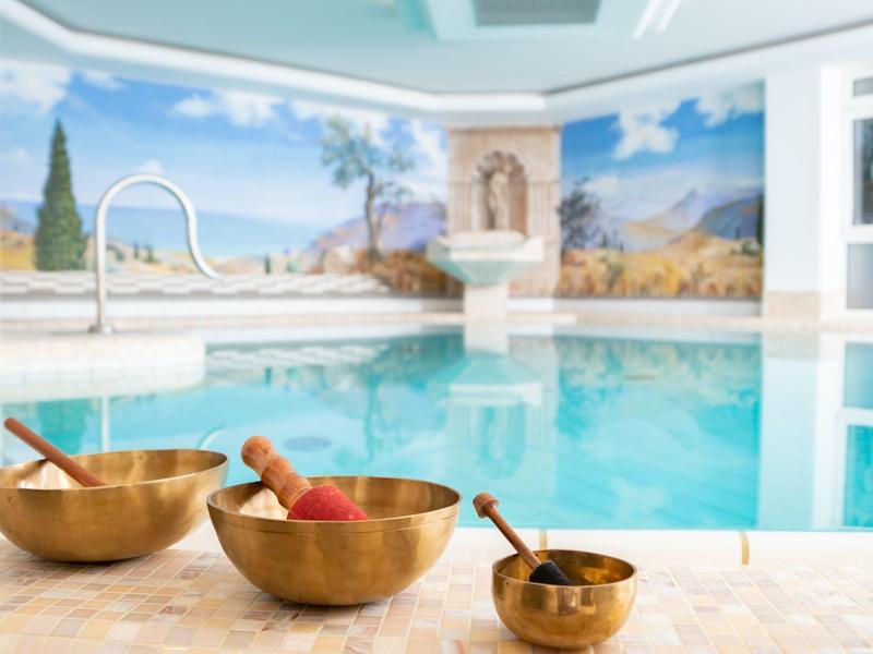 Una piscina coperta con acqua turchese, murales dipinti e tre ciotole sonore in primo piano.