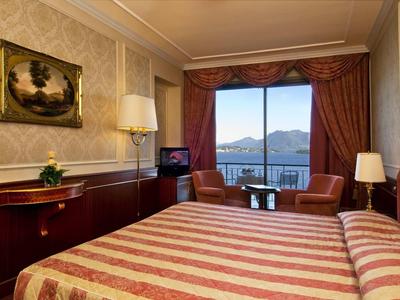 Hotelzimmer mit Doppelbett, roten Vorhängen und Blick auf Berge und See durch großes Fenster.