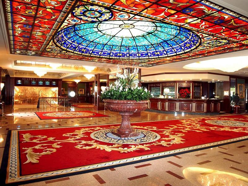 Hotel lobby met gekleurde glasplafond, grote rode tapijten en bloemstuk in het midden.