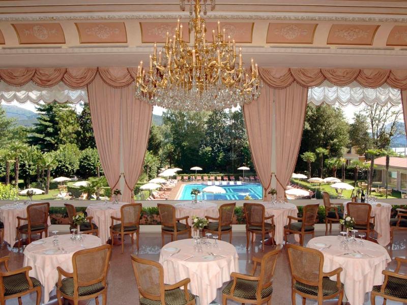 Bright terrace with round tables, chairs, chandelier, and view of pool and garden.