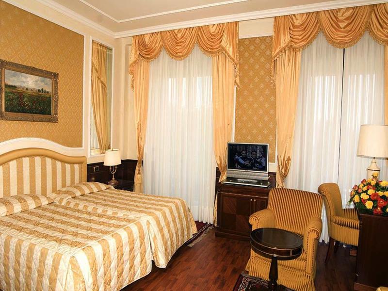 Elegant hotel room with double bed, armchair, desk, and classic gold-toned decor.