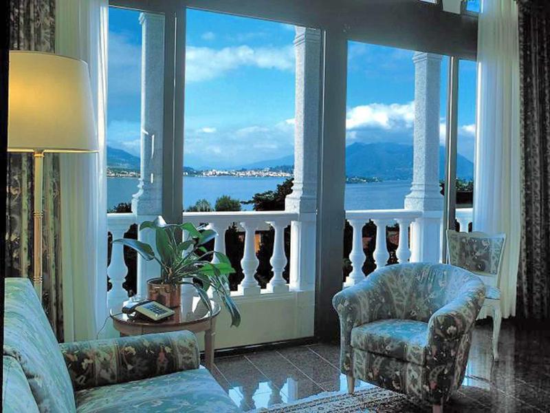 View from a room with floral armchairs onto a balcony and lake with mountains in the background.