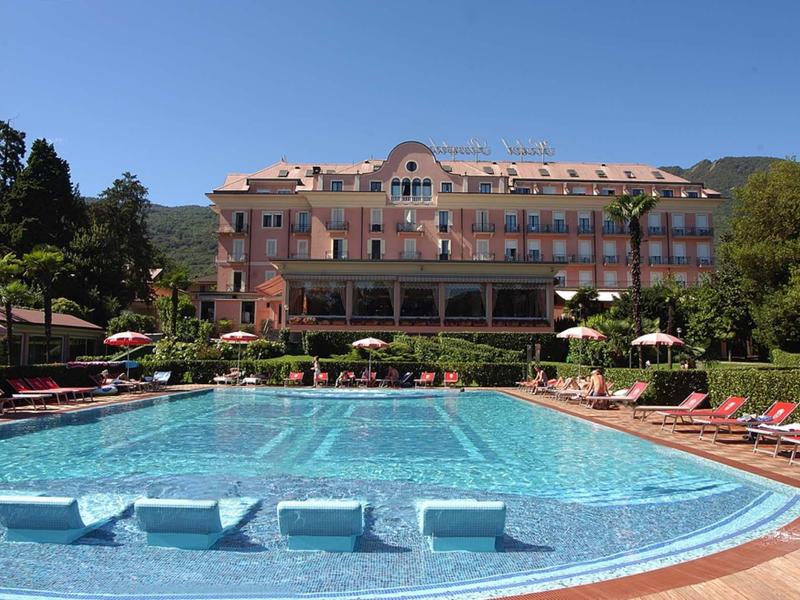 Large outdoor pool with sun loungers in front of a multi-story hotel building and trees.
