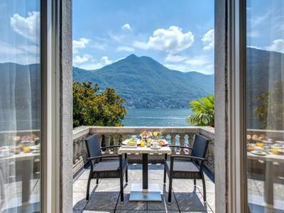 Vista attraverso porte aperte su balcone con tavolo e sedie, montagna e lago sullo sfondo.