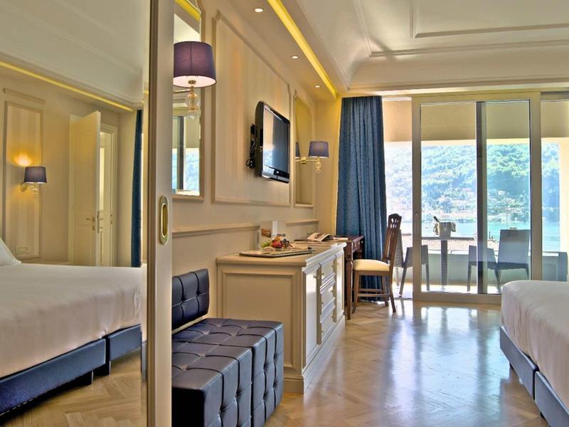 Camera d'albergo luminosa con due letti, TV e balcone con vista sul mare.