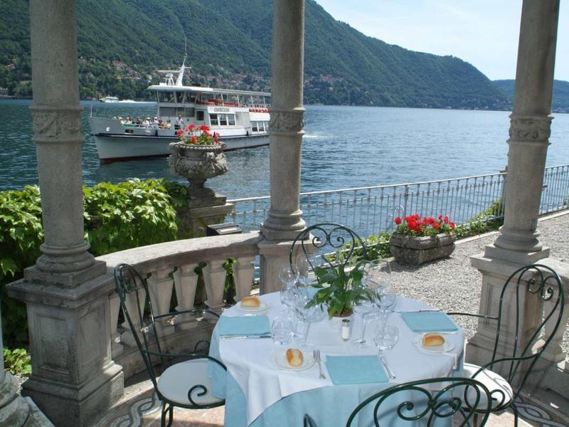 Terrazza con tavolo e sedie sul lago, con una barca e montagne sullo sfondo.