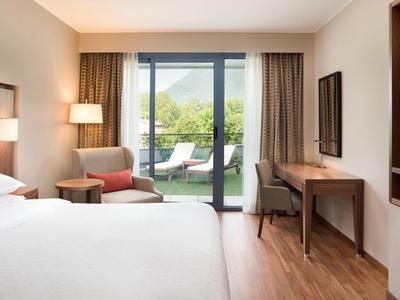 Modernes Hotelzimmer mit großem Bett, Sessel, Schreibtisch und Blick auf Terrasse und Berge.