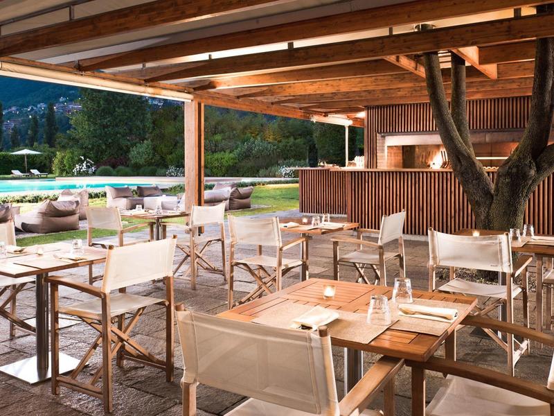 Open eetgedeelte met houten tafels en stoelen onder een houten constructie met uitzicht op het zwembad en de bergen.