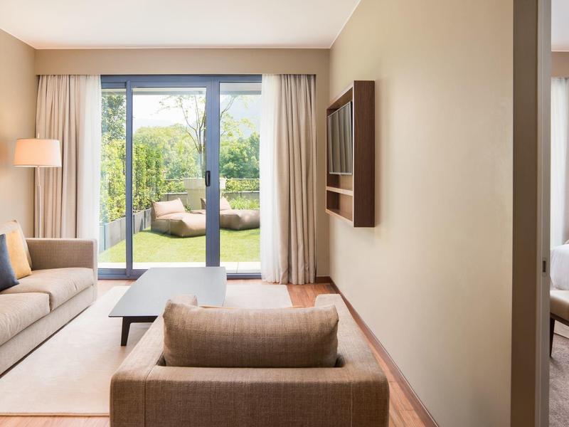 Modern ingericht hotelkamer met woonkamer, slaapkamer en uitzicht op tuin door grote ramen.