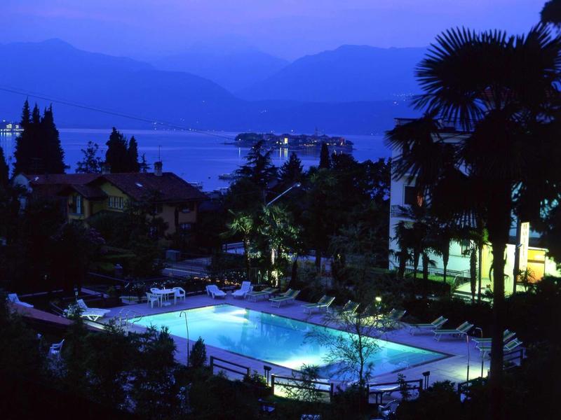 Piscina illuminata in un hotel con montagne e lago sullo sfondo di notte.
