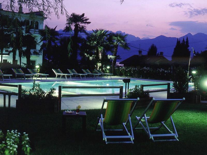 Piscina illuminata con sedie a sdraio in giardino di un hotel al crepuscolo con montagne sullo sfondo.