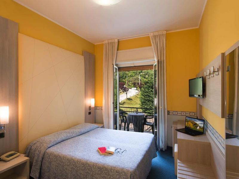 Piccola camera d'hotel con letto matrimoniale, pareti gialle e balcone con vista verde.