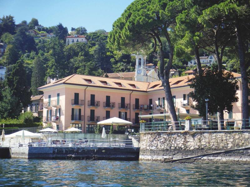 Hotel aan een meer met terrassen en bomen op een zonnige dag.