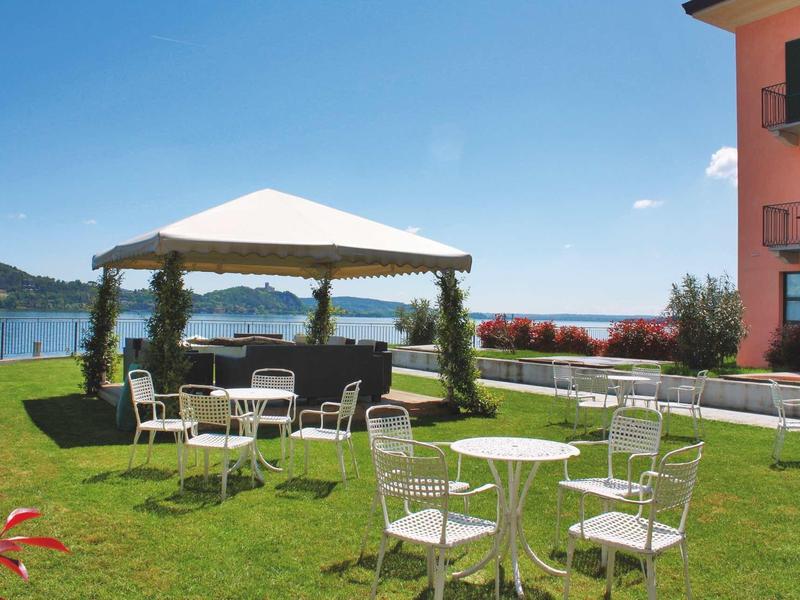 Tuin met witte stoelen en tafels onder een prieel naast een rood hotelgebouw aan het meer.