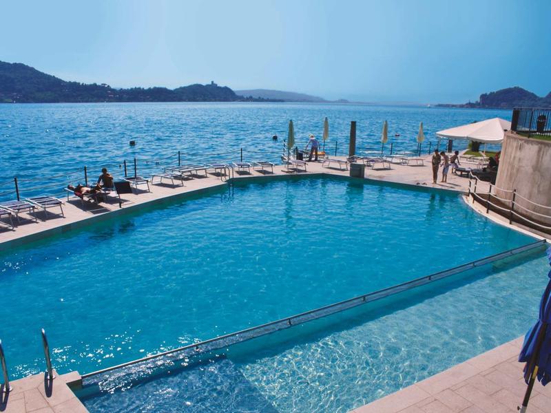 Groot buitenzwembad met ligstoelen aan de kust en uitzicht op bergen bij zonnig weer.