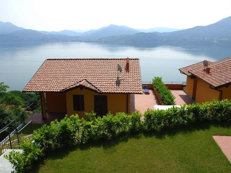 Haus mit rotem Ziegeldach, grünem Garten und Blick auf ruhigen See mit Bergen im Hintergrund.