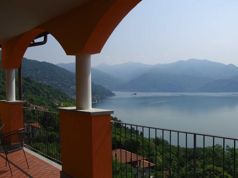 Blick auf einen See mit Bergen im Hintergrund, Terrasse mit orangefarbenen Säulen und Stühlen im Vordergrund.