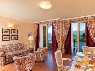 Gemütliches Hotelzimmer mit floral gemusterten Vorhängen und Sesseln sowie Blick auf See und Berge.