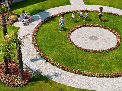 Runder Garten mit grünem Rasen, Blumenrabatten und einem gepflasterten Weg, Familien spazieren.