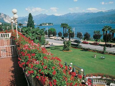 Vue d'un balcon avec des fleurs rouges sur un parc et un lac avec des montagnes sous un ciel ensoleillé.