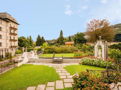Grüner Park mit Wegen, Bänken, Blumenbeeten und einem mehrstöckigen Gebäude links.
