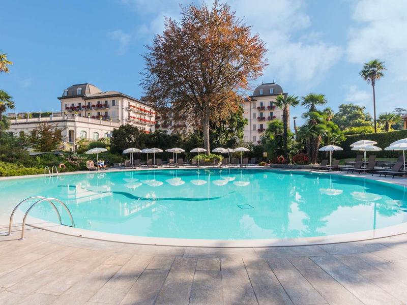 Grande piscine d'hôtel avec parasols et palmiers, entourée d'un bâtiment et ciel bleu.