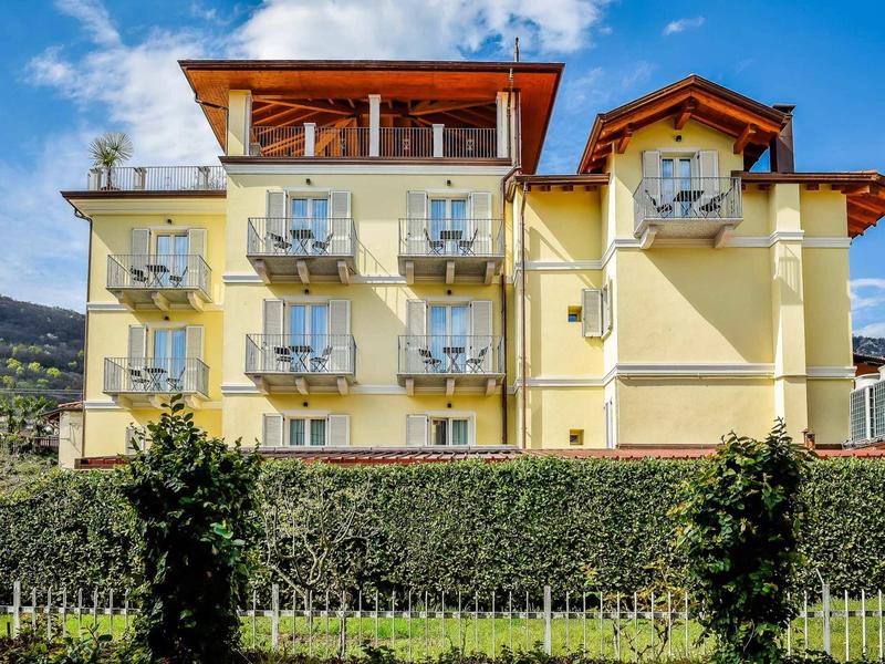 Mehrstöckiges gelbes Gebäude mit Balkonen, umgeben von grünem Hecken und Bäumen, klarer Himmel.